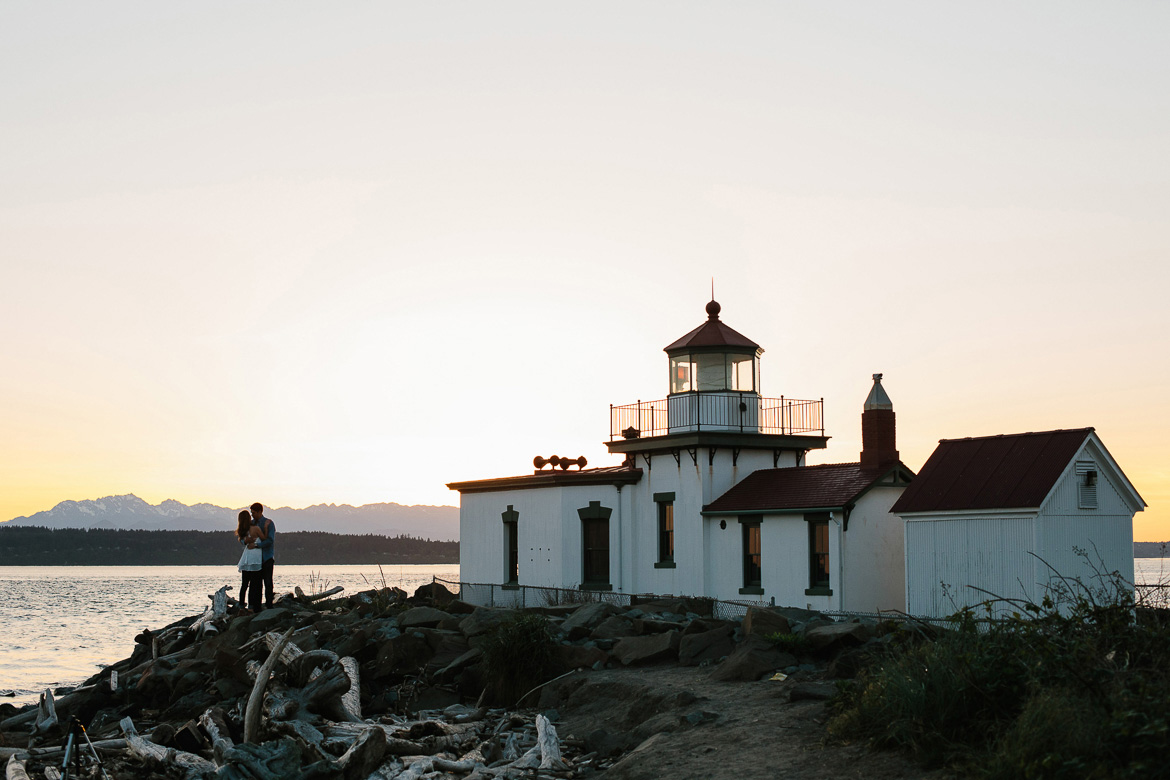 Couple on rocky beach by lighthouse during sunset engagement photos at Discovery Park in Seattle, WA