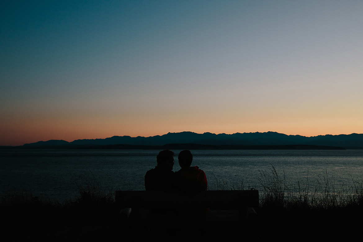Couple sitting on beach during Whidbey Island engagement session looking at the Olympic Mountians